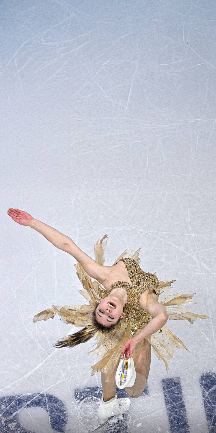 A high-angle shot of figure skater Alysa Liu performing a dynamic spin on the ice in a gold feathered dress. This sports wallpaper captures the elegance and motion of professional skating for your iPhone. | sports athlete olympics iphone mobile lock screen 4k wallpaper hd wallpaper home screen winter sports ice skating gold dress static wallpaper alysa liu figure skating ice rink figure skater sports wallpapers A high-angle shot of figure skater Alysa Liu performing a dynamic spin on the ice in a gold feathered dress. This sports wallpaper captures the elegance and motion of professional skating for your iPhone.