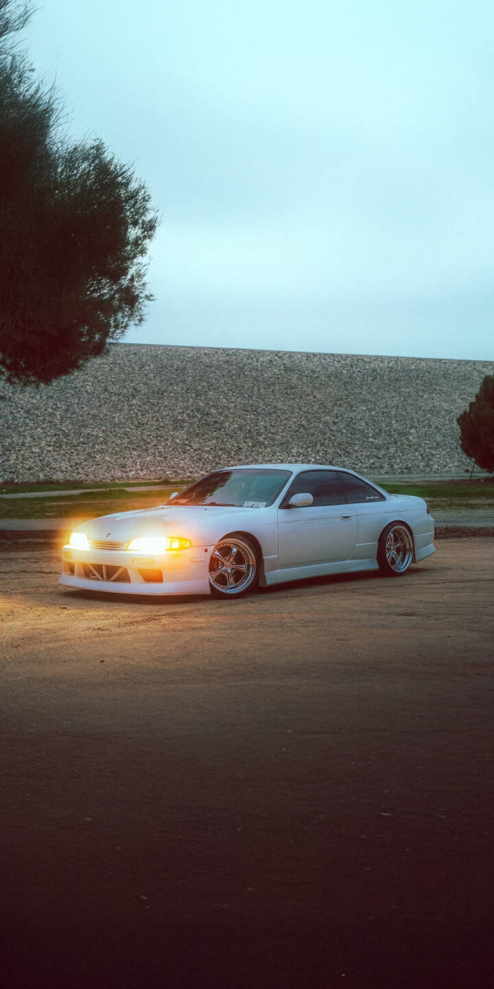 Download this stunning Nissan Silvia S14 4K wallpaper featuring a pristine white car with bright, glowing headlights and gleaming chrome wheels. This high-resolution 4K wallpaper is perfect for personalizing your iPhone, showcasing the iconic car against a textured background. | sports car iphone nissan mobile white car 4k wallpaper hd wallpaper car wallpaper jdm modified car nissan silvia s14 s14 Download this stunning Nissan Silvia S14 4K wallpaper featuring a pristine white car with bright, glowing headlights and gleaming chrome wheels. This high-resolution 4K wallpaper is perfect for personalizing your iPhone, showcasing the iconic car against a textured background.