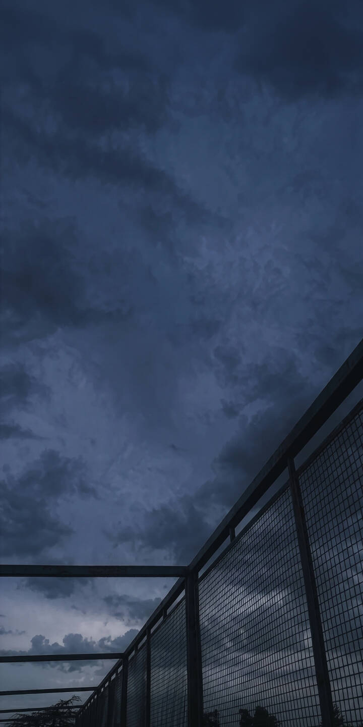 Heavy, foreboding dark blue clouds dominate the sky, creating an ominous mood over a striking geometric metal fence in this compelling 4K wallpaper. The contrast of the somber sky and industrial foreground makes this a unique atmospheric wallpaper perfect for iPhone. | urban iphone clouds wallpaper mobile 4k wallpaper hd wallpaper dark aesthetic gloomy stormy sky atmospheric metal fence Heavy, foreboding dark blue clouds dominate the sky, creating an ominous mood over a striking geometric metal fence in this compelling 4K wallpaper. The contrast of the somber sky and industrial foreground makes this a unique atmospheric wallpaper perfect for iPhone.