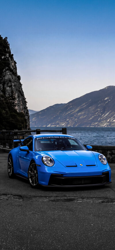 A vibrant blue Porsche 911 GT3 sports car is perfectly set against a stunning lake and rugged mountain landscape in this 4K iPhone wallpaper. This matching wallpaper, ideal for two screens, showcases the elegant car and picturesque scenery, making it a captivating mobile wallpaper.