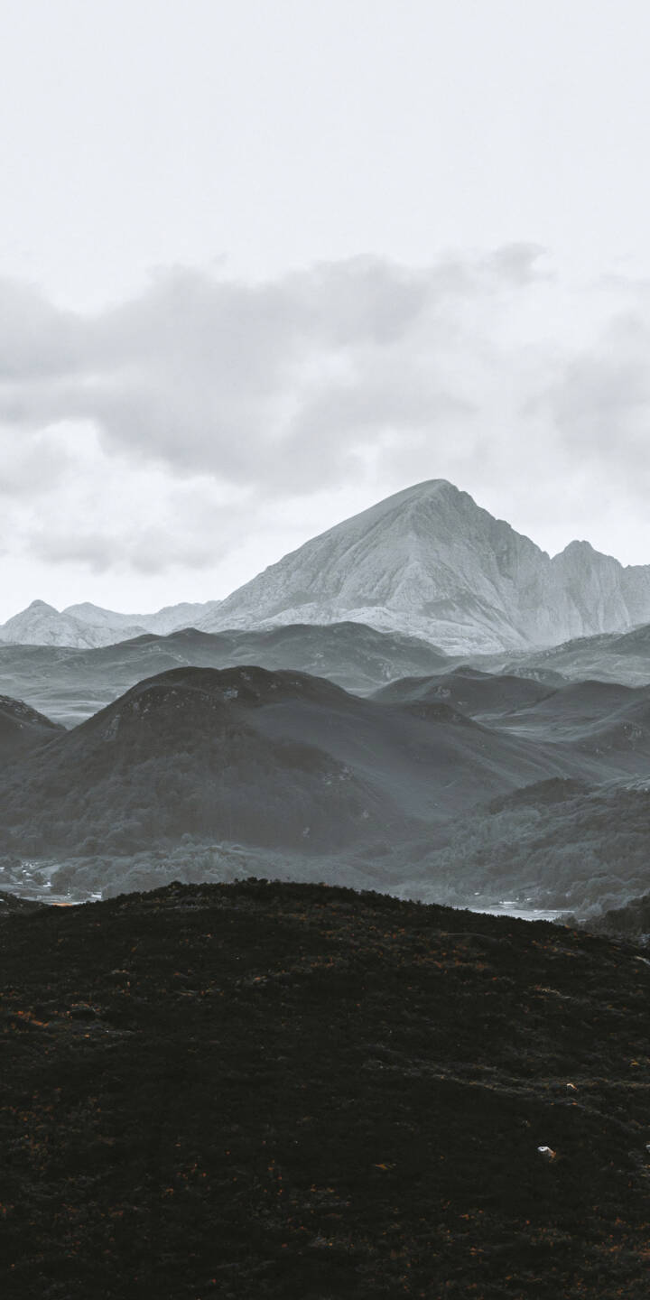 A majestic mountain peak rises above layers of rugged hills beneath a vast, overcast sky. Soft gray clouds drift across the distant summit, while dark foreground ridges gradually fade into misty mountain valleys. The restrained monochromatic palette, natural textures, and dramatic contrast between light and shadow create a timeless cinematic landscape filled with solitude, tranquility, and the untouched beauty of the wilderness. | landscape nature mountains monochrome wilderness A majestic mountain peak rises above layers of rugged hills beneath a vast, overcast sky. Soft gray clouds drift across the distant summit, while dark foreground ridges gradually fade into misty mountain valleys. The restrained monochromatic palette, natural textures, and dramatic contrast between light and shadow create a timeless cinematic landscape filled with solitude, tranquility, and the untouched beauty of the wilderness.