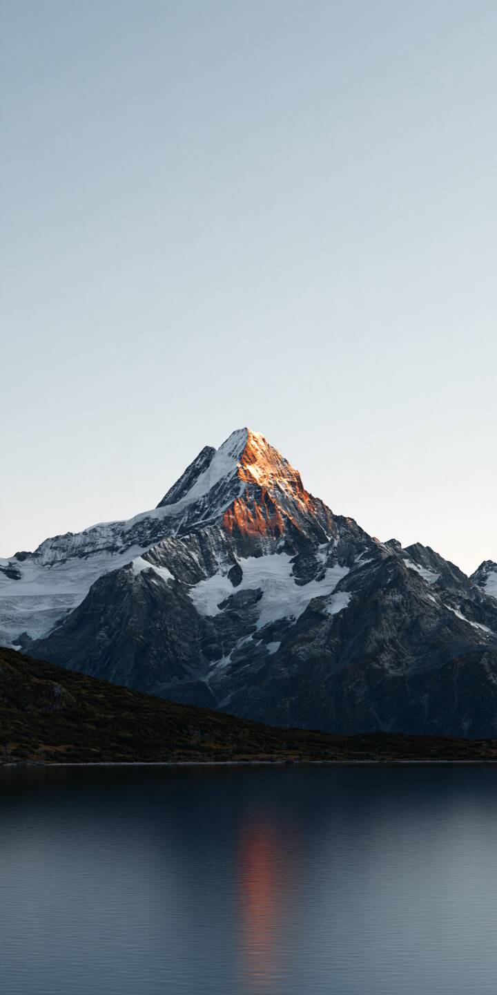 A majestic snow-covered mountain rises above a peaceful alpine lake, its rugged summit illuminated by the warm golden light of the rising or setting sun. The glowing orange highlights contrast beautifully with the cool blue-gray mountain slopes, while a delicate reflection stretches across the still water below. Beneath a vast, pale sky, the minimalist composition captures the breathtaking beauty of the wilderness, creating a timeless atmosphere of tranquility, solitude, and natural grandeur. | lake snow sunrise reflection alpine A majestic snow-covered mountain rises above a peaceful alpine lake, its rugged summit illuminated by the warm golden light of the rising or setting sun. The glowing orange highlights contrast beautifully with the cool blue-gray mountain slopes, while a delicate reflection stretches across the still water below. Beneath a vast, pale sky, the minimalist composition captures the breathtaking beauty of the wilderness, creating a timeless atmosphere of tranquility, solitude, and natural grandeur.
