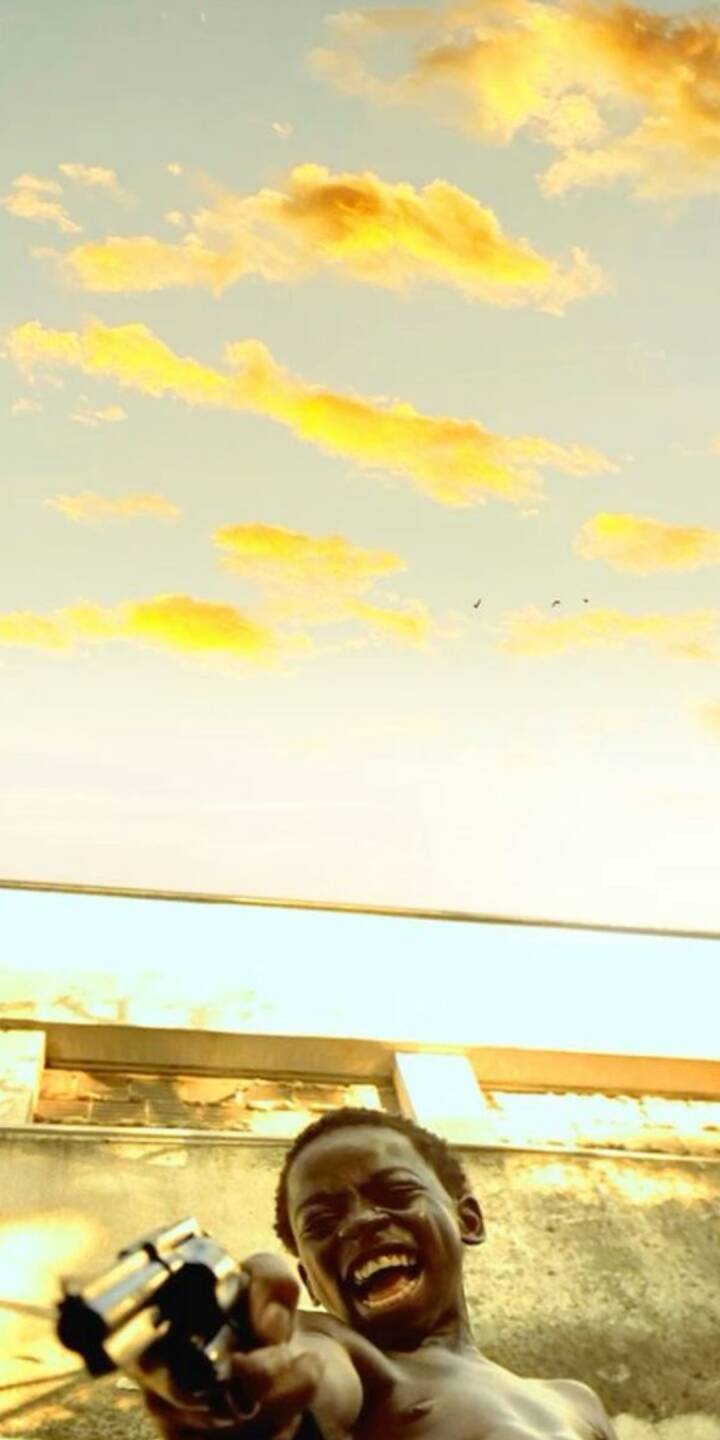 A chilling, maniacal grin stretches across the young Lil' Dice's face as he points a gleaming revolver directly at the viewer, bathed in the warm, golden light of a dramatic sky. This powerful 4K wallpaper masterfully captures the raw intensity of the iconic City of God character, making it a gripping and unforgettable iPhone wallpaper. | artist A chilling, maniacal grin stretches across the young Lil' Dice's face as he points a gleaming revolver directly at the viewer, bathed in the warm, golden light of a dramatic sky. This powerful 4K wallpaper masterfully captures the raw intensity of the iconic City of God character, making it a gripping and unforgettable iPhone wallpaper.