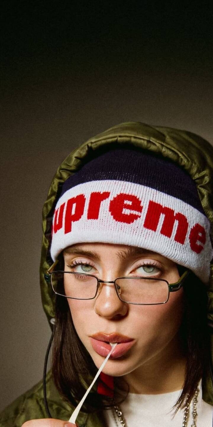 Billie Eilish's iconic green eyes look up as she subtly pulls a string of bubblegum, featuring her stylish Supreme beanie and glasses in this striking 4K wallpaper – a perfect wallpaper for iPhone. | artist Billie Eilish's iconic green eyes look up as she subtly pulls a string of bubblegum, featuring her stylish Supreme beanie and glasses in this striking 4K wallpaper – a perfect wallpaper for iPhone.
