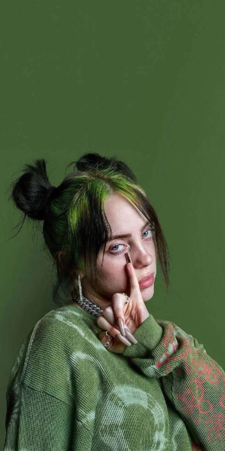 Billie Eilish, with her signature green hair streaks and double buns, presents an enigmatic expression as she delicately touches her finger beneath her eye, making for a uniquely aesthetic 4K wallpaper. This captivating green-themed image is a perfect mobile wallpaper for any iPhone user looking for an artistic celebrity wallpaper. | artist Billie Eilish, with her signature green hair streaks and double buns, presents an enigmatic expression as she delicately touches her finger beneath her eye, making for a uniquely aesthetic 4K wallpaper. This captivating green-themed image is a perfect mobile wallpaper for any iPhone user looking for an artistic celebrity wallpaper.