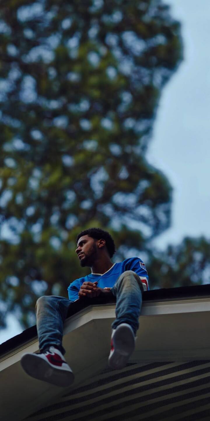 J. Cole sits thoughtfully on a house eave, wearing a blue jersey and jeans with red-accented sneakers, as blurred trees provide a natural backdrop in this serene 4K wallpaper. This contemplative J. Cole image makes an excellent wallpaper for iPhone users. | artist J. Cole sits thoughtfully on a house eave, wearing a blue jersey and jeans with red-accented sneakers, as blurred trees provide a natural backdrop in this serene 4K wallpaper. This contemplative J. Cole image makes an excellent wallpaper for iPhone users.