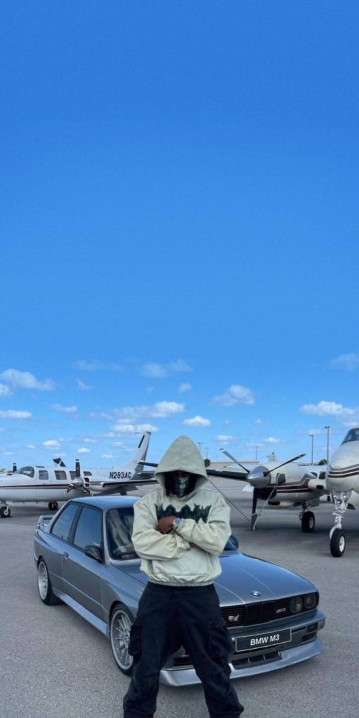 A mysterious masked figure in a light-colored hoodie stands coolly with crossed arms in front of a gleaming silver BMW M3, backdropped by private jets under a wide blue sky in this high-definition 4K wallpaper. This stylish airport scene makes an excellent wallpaper for iPhone users seeking a unique urban aesthetic. | artist A mysterious masked figure in a light-colored hoodie stands coolly with crossed arms in front of a gleaming silver BMW M3, backdropped by private jets under a wide blue sky in this high-definition 4K wallpaper. This stylish airport scene makes an excellent wallpaper for iPhone users seeking a unique urban aesthetic.